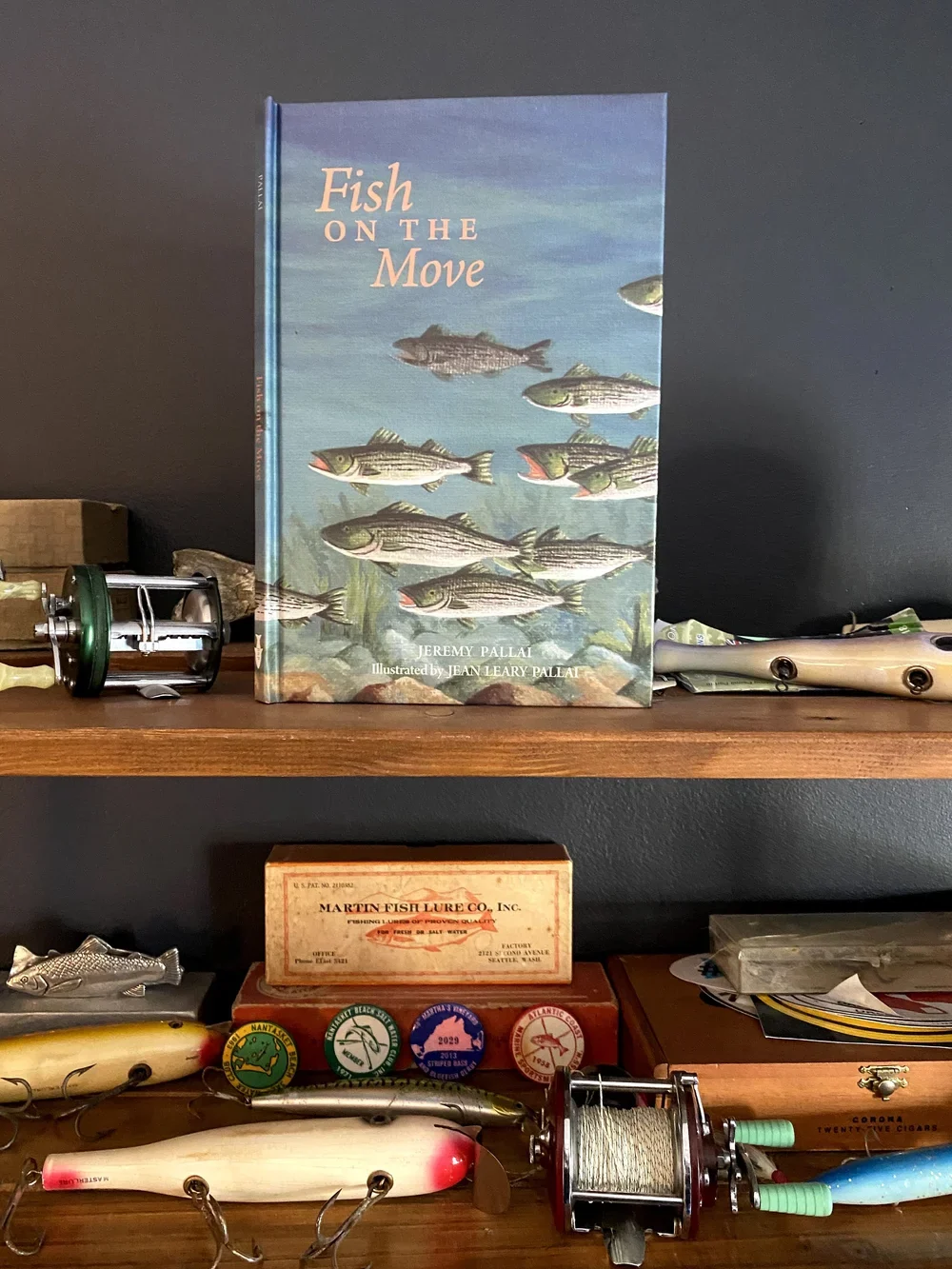 The hardcover of Fish on the Move standing on a shelf among vintage fishing reels, plugs and striped bass club badges.